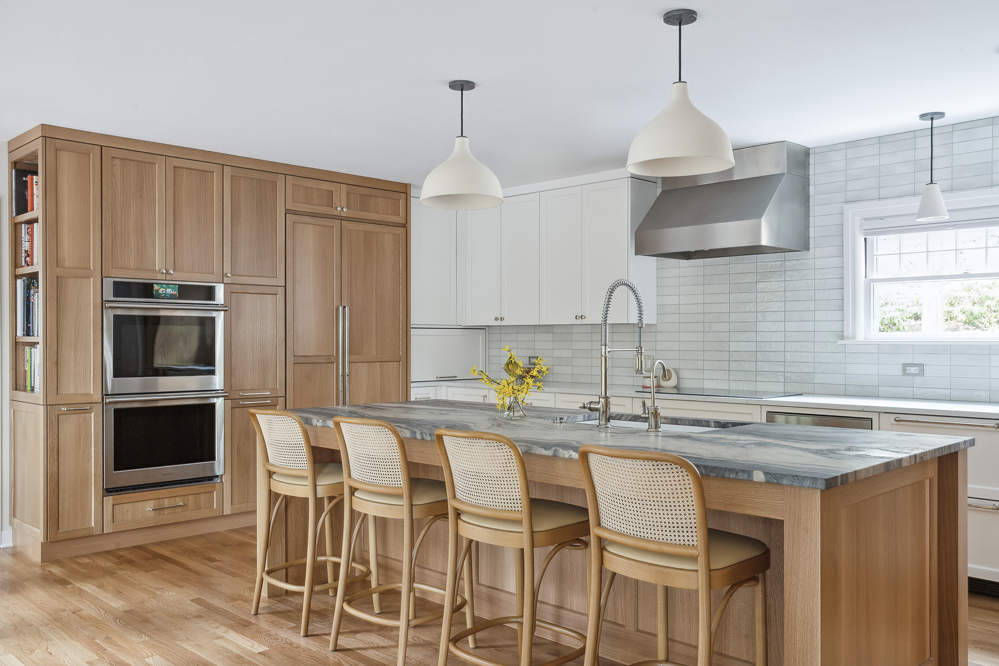 Remodeled kitchen showing oak storage wall with double ovens and bookshelf beside island with cane barstools, Arlington Heights Remodel by Tweto Architecture
