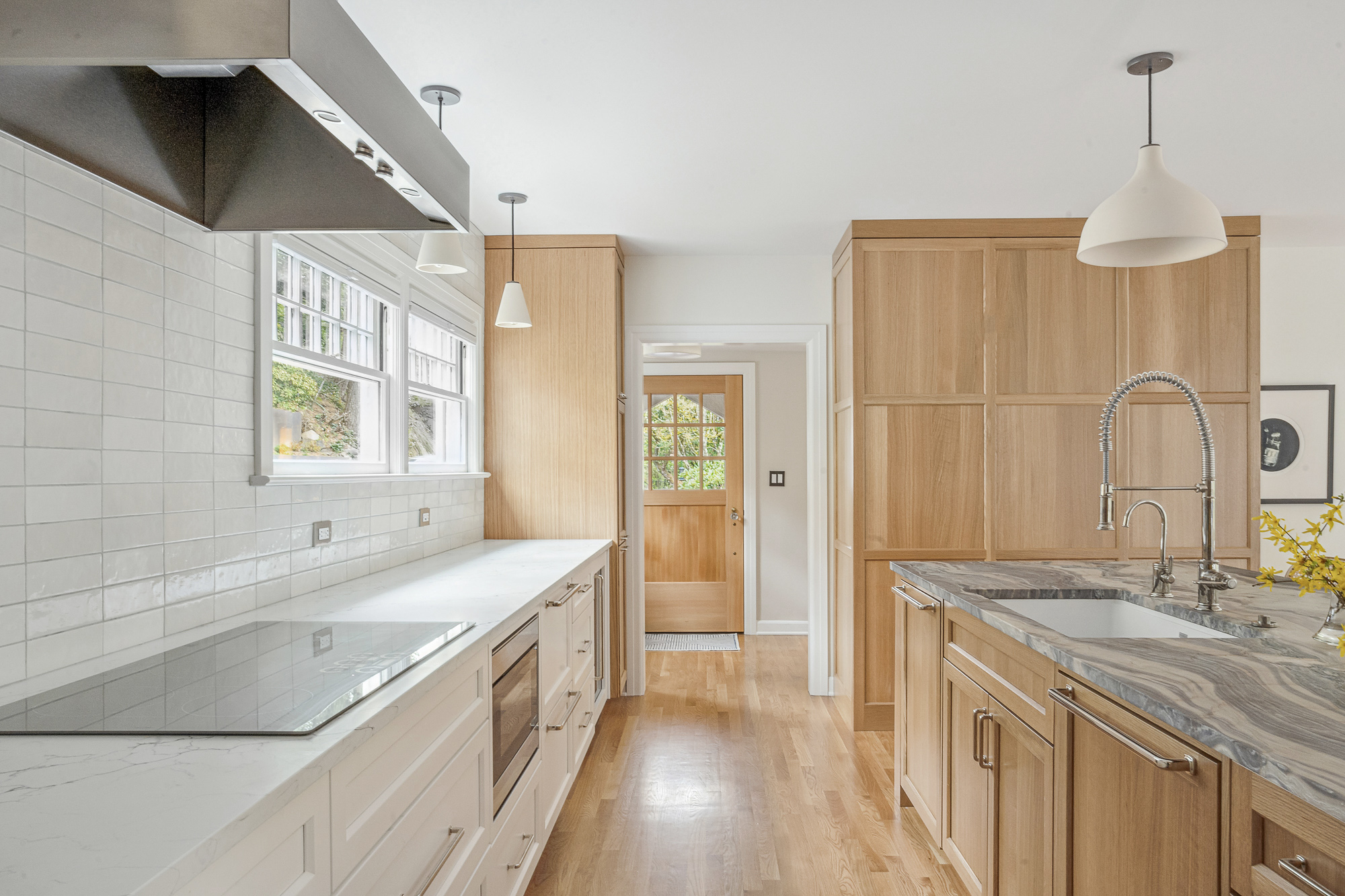 Kitchen looking toward exterior door with arched window, white tile backsplash and induction cooktop at left, oak storage wall at right, Arlington Heights Remodel by Tweto Architecture