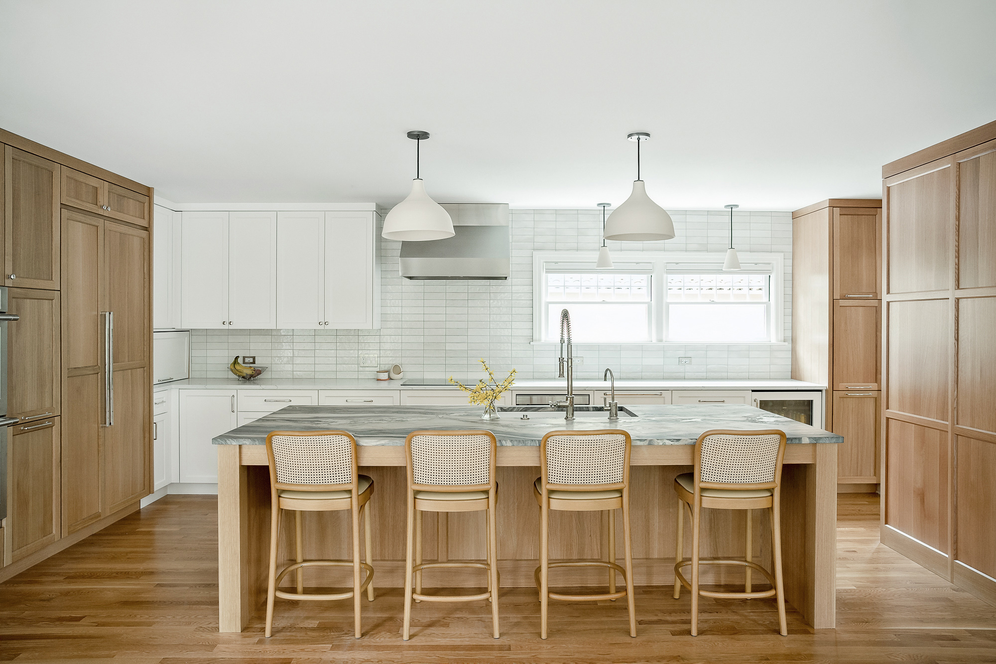 Kitchen island from the front with four cane barstools, white perimeter cabinets and oak storage wall beyond, Arlington Heights Remodel by Tweto Architecture