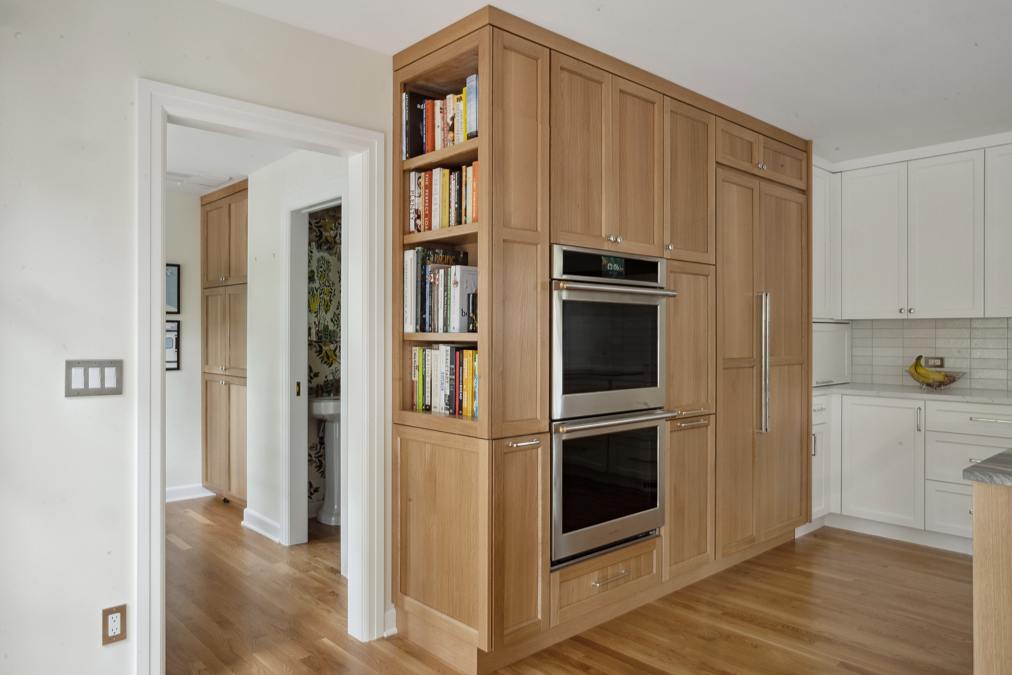 Oak storage wall with open cookbook shelving flanking double wall ovens, doorway to hall and half bath visible at left, Arlington Heights Remodel by Tweto Architecture
