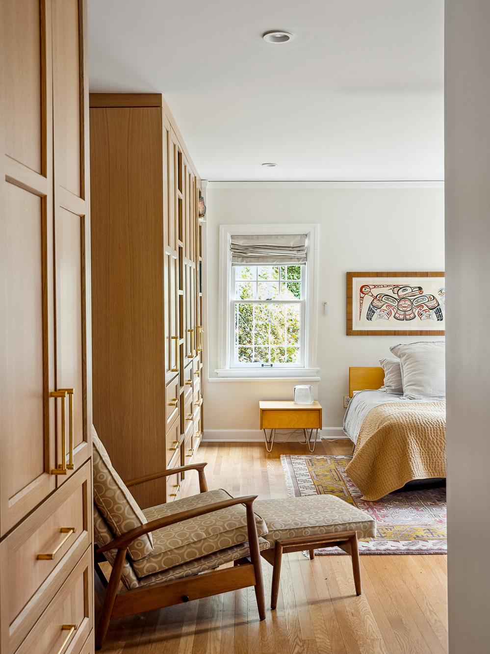 Primary bedroom with tall oak built-in wardrobe and reading chair beside bed, view to window with roman shade, Arlington Heights Remodel by Tweto Architecture