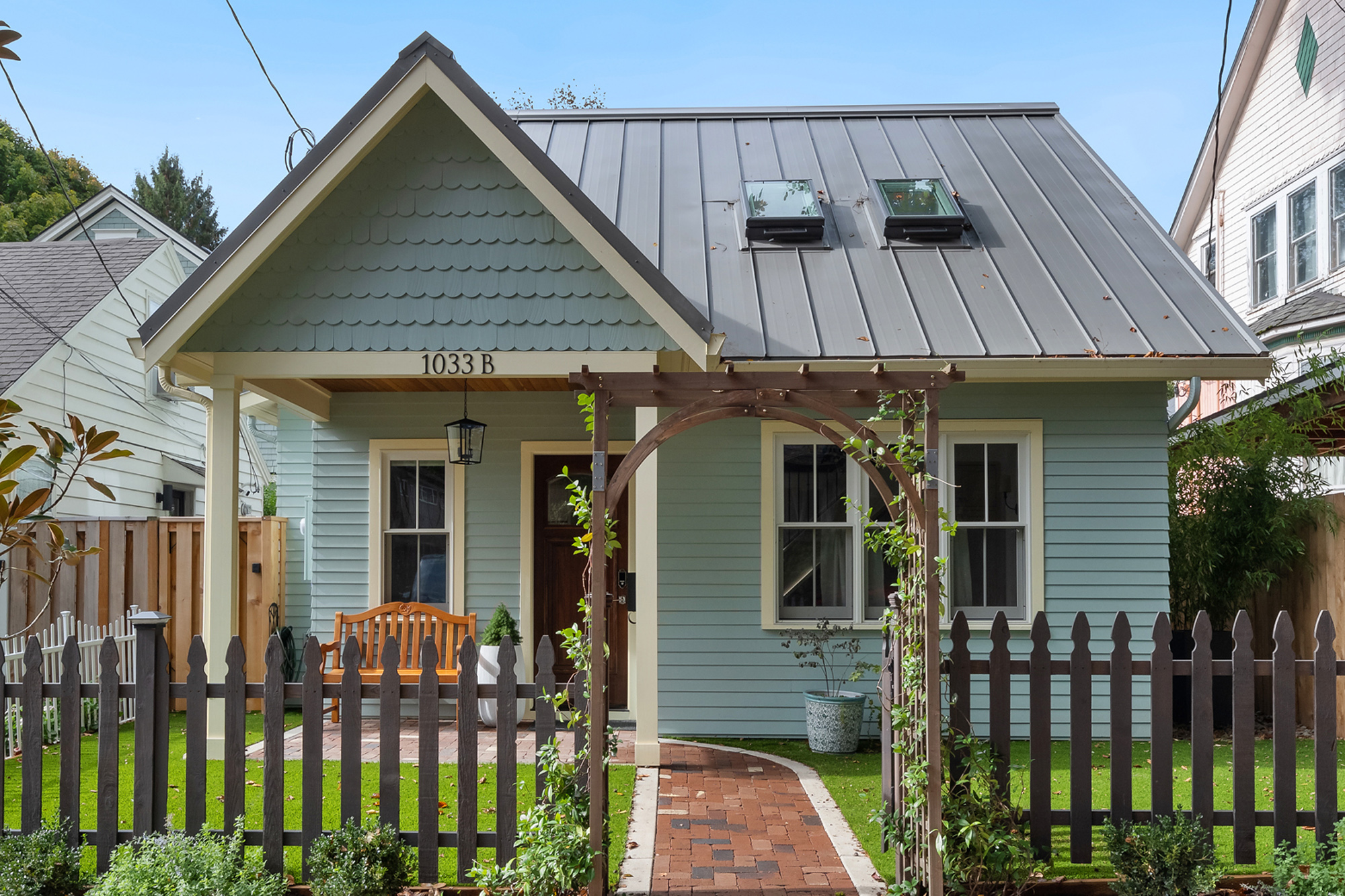 Buckman ADU front exterior, a pale green cottage with metal roof, picket fence, brick path, and arbor trellis at the entry, SE Portland by Tweto Architecture