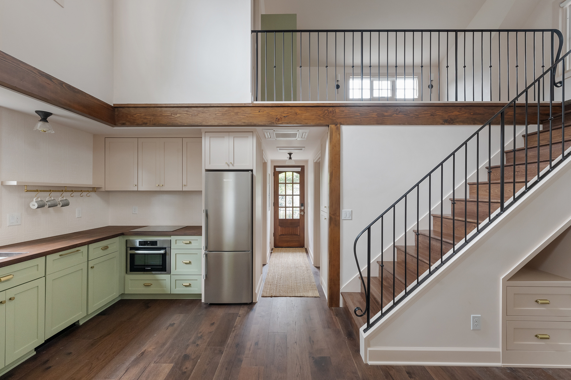 Kitchen with green cabinets and walnut countertop at left, stair with painted steel railing at right, arched front door visible at the end of the hall, Buckman ADU SE Portland by Tweto Architecture
