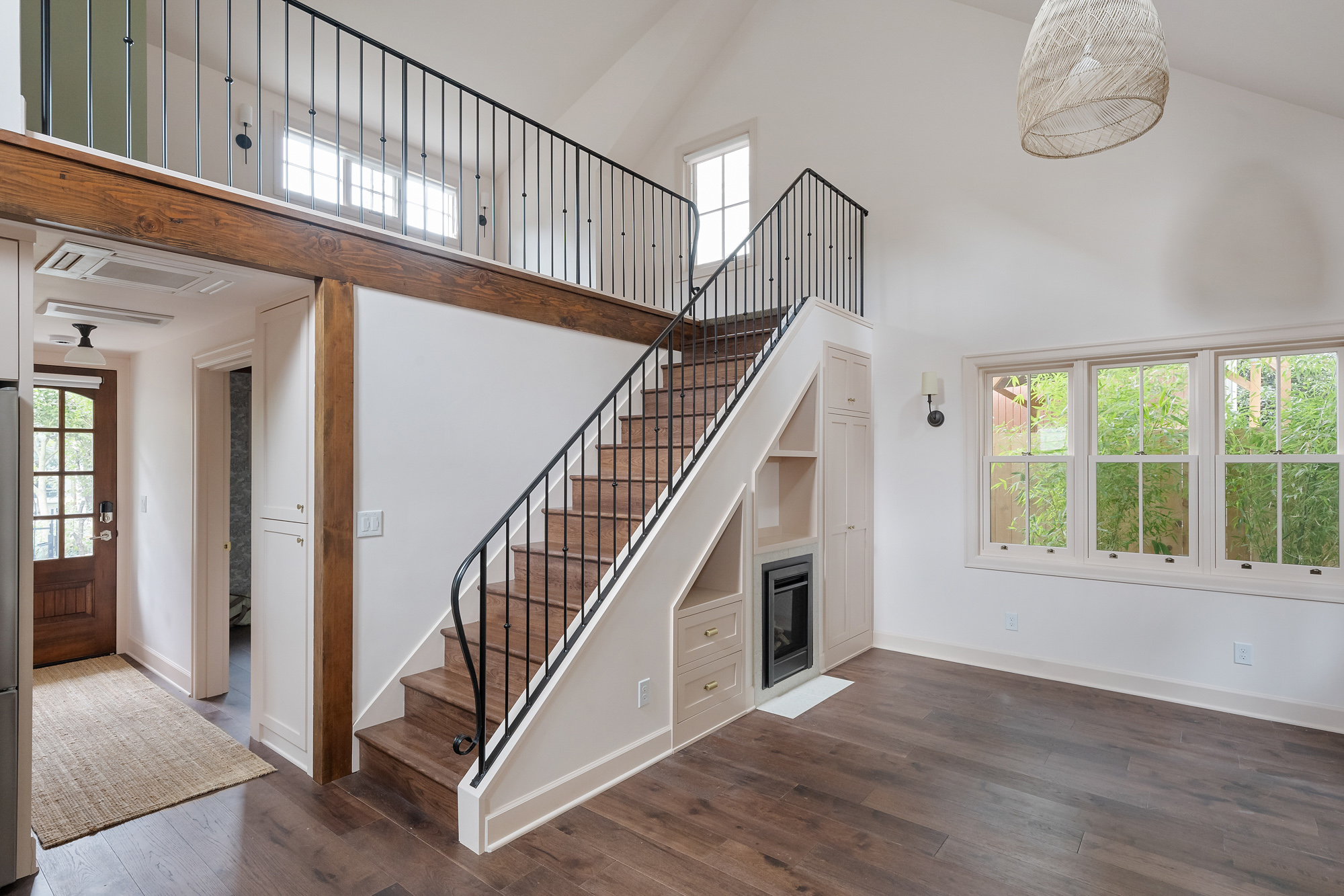 Stair with painted steel railing and built-in storage and fireplace below, open living area with tall windows at right, Buckman ADU SE Portland by Tweto Architecture