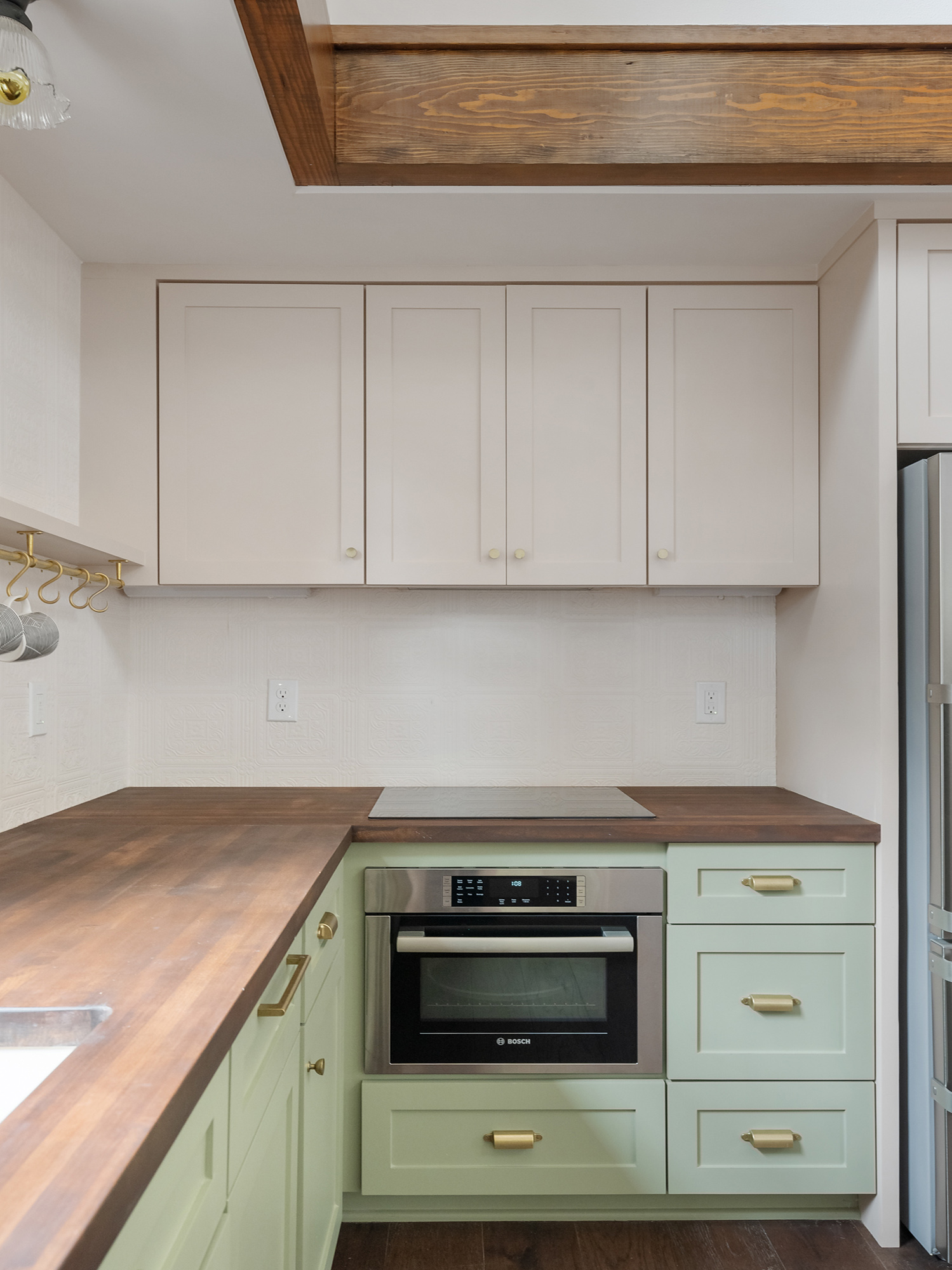 Kitchen range wall with green lower cabinets, cream upper cabinets, walnut countertop, and Bosch wall oven beneath an exposed wood beam, Buckman ADU SE Portland by Tweto Architecture