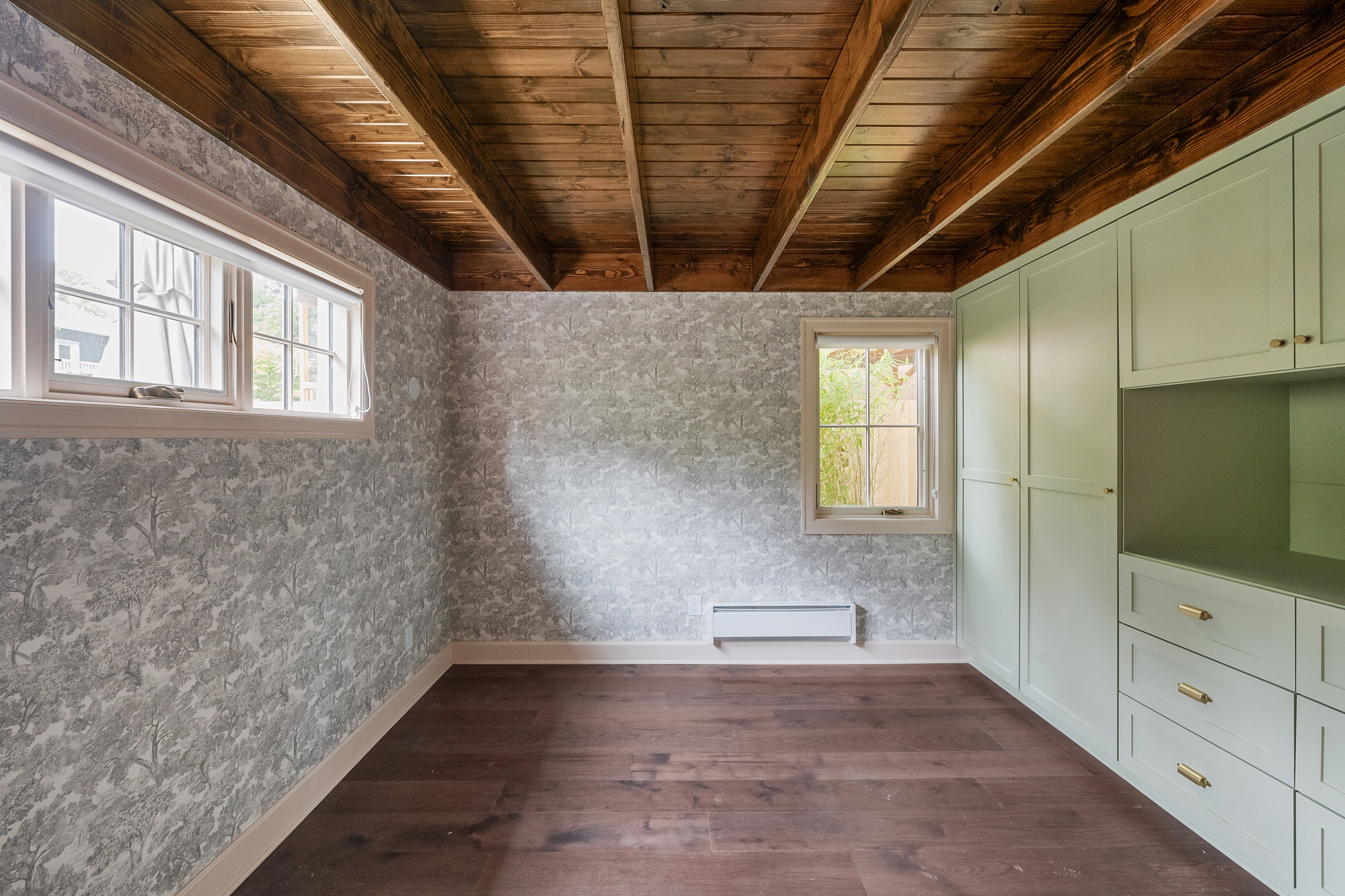 Main-floor bedroom with exposed wood beam ceiling, grey toile wallpaper, built-in green wardrobe, and windows to the side yard, Buckman ADU SE Portland by Tweto Architecture