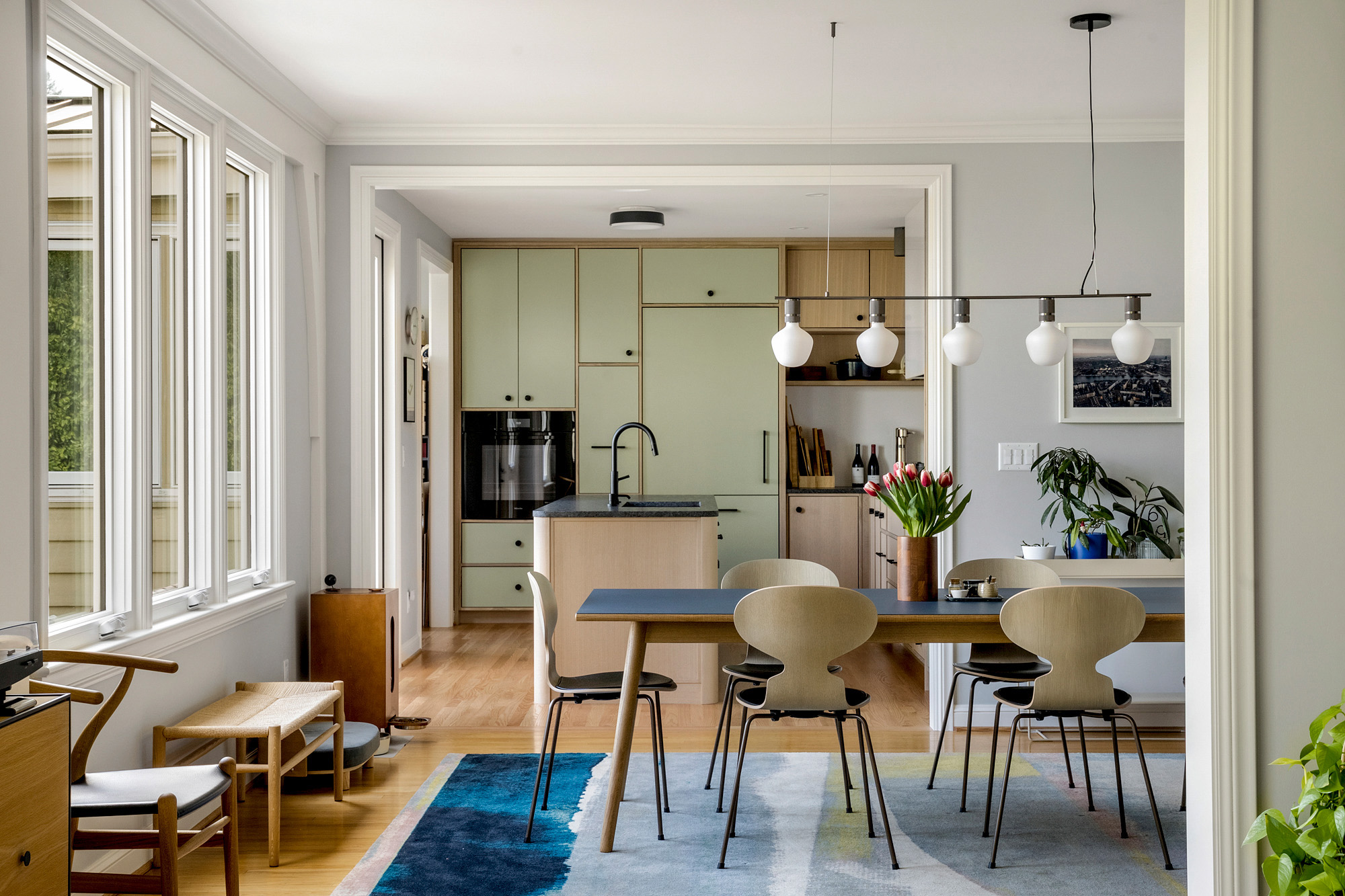 View from art studio nook across dining table toward sage green kitchen with island beyond, by Tweto Architecture