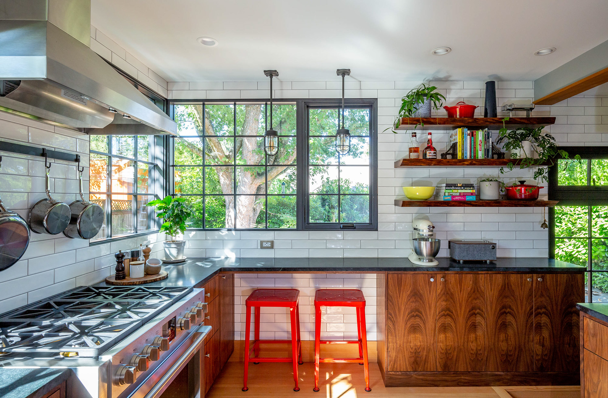 Cooking side of the kitchen with commercial range, red metal bar stools at the soapstone seating counter, and black-framed windows overlooking the yard, Concordia Kitchen by Tweto Architecture