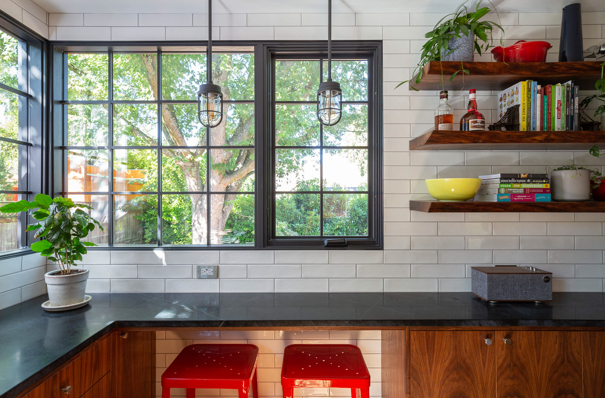 Seating counter with soapstone top and red bar stools beneath open walnut shelves and tall black-framed windows, Concordia Kitchen by Tweto Architecture