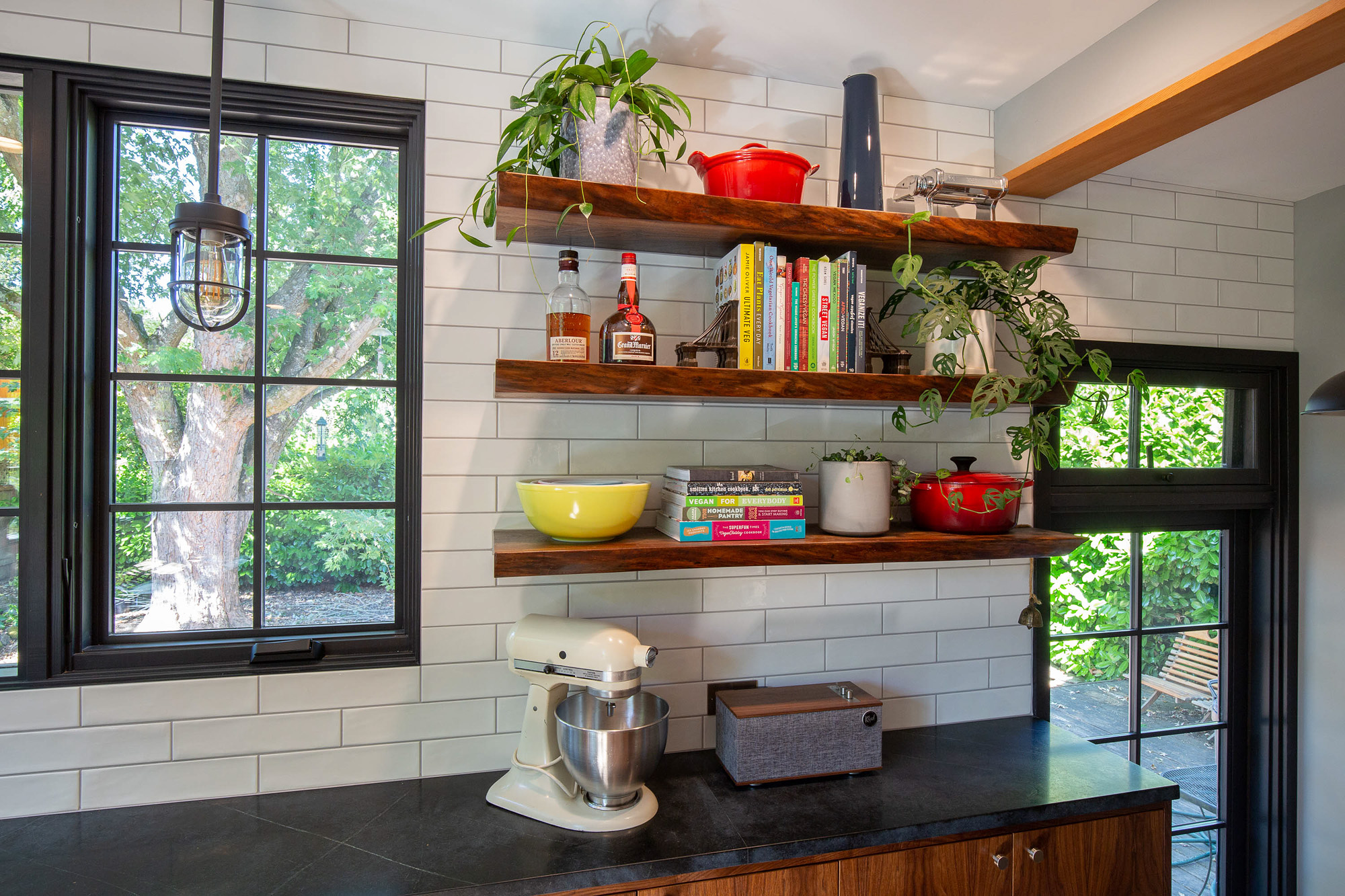 Three open walnut floating shelves loaded with cookbooks and plants against full-height white subway tile, with stand mixer on the soapstone counter below, Concordia Kitchen by Tweto Architecture