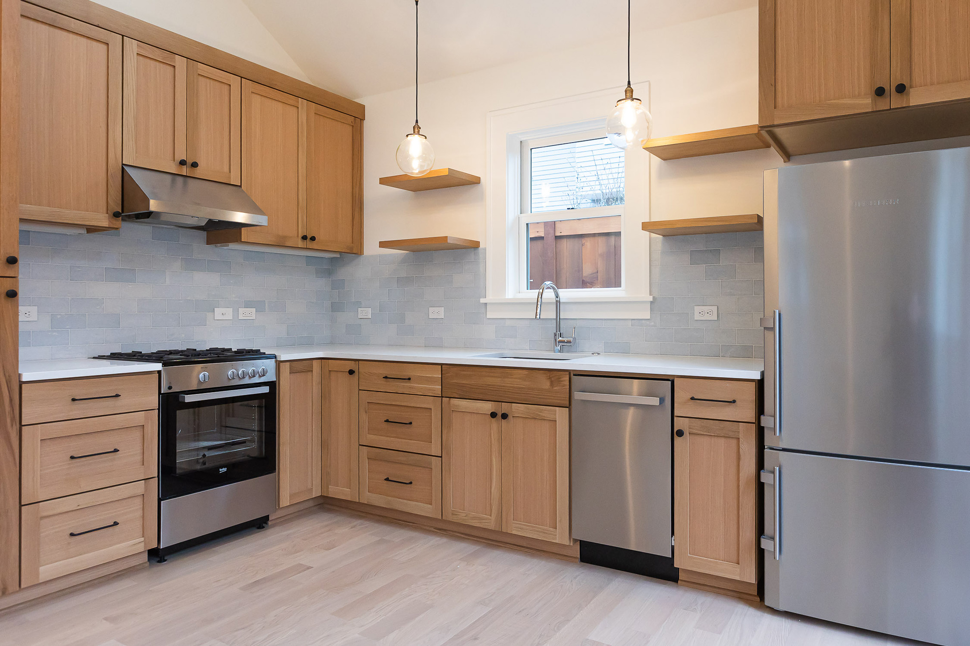 Kitchen with white oak shaker cabinets, grey subway tile backsplash, floating wood shelves, globe pendants, and stainless appliances, Irvington ADU by Tweto Architecture