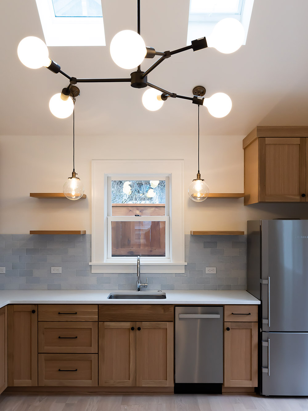 Kitchen sink wall with skylights above, globe chandelier, grey subway tile backsplash, floating oak shelves, and stainless appliances, Irvington ADU by Tweto Architecture