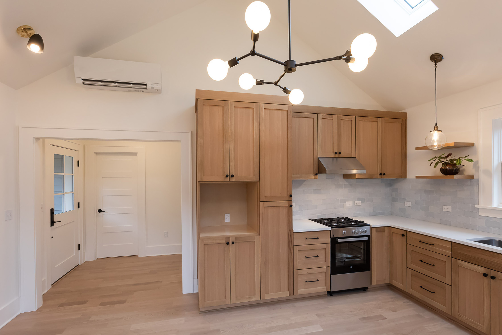 Great room and kitchen with vaulted ceiling, tall oak storage wall, gas range, globe pendant chandelier, mini-split, and entry door beyond, Irvington ADU by Tweto Architecture