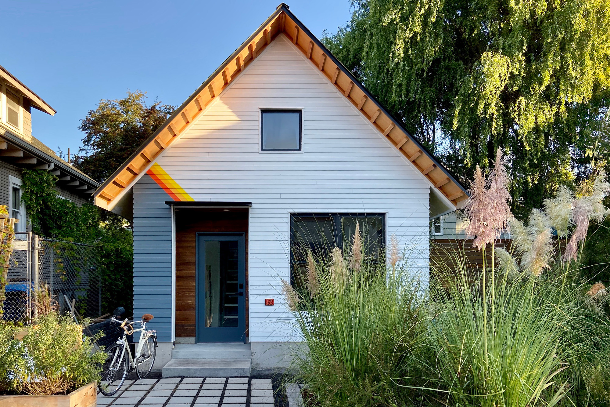 Mason Street ADU front elevation, white gable with exposed fir eave and wood-clad recessed entry, ornamental grasses and a bicycle by the door, NE Portland, by Tweto Architecture