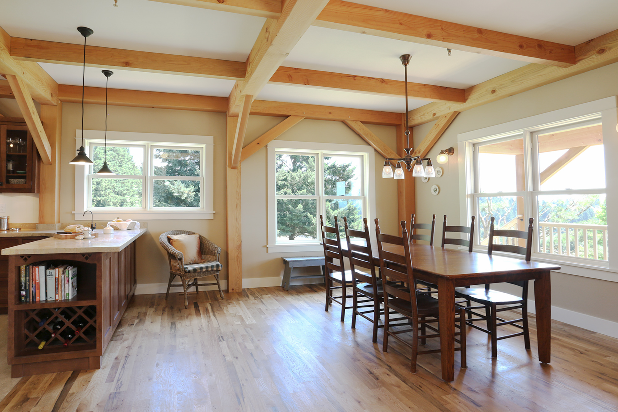 Dining room with exposed Douglas Fir timber frame, farmhouse table, and south-east windows, kitchen peninsula with wine rack and cookbook storage to left, North Plains Timber Frame by Tweto Architecture