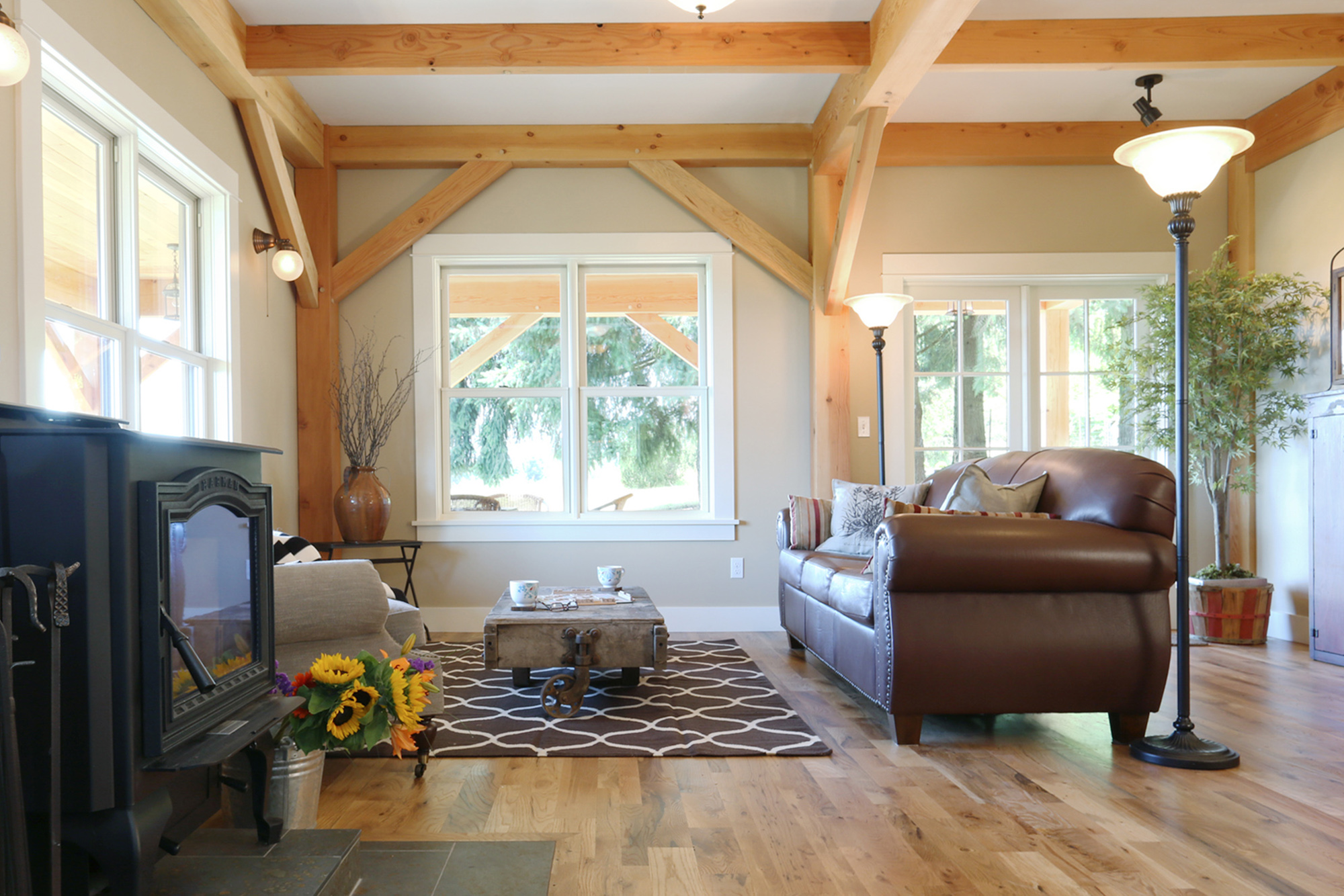 Living room with exposed timber knee brace, wood stove, leather sofa, and south-facing windows, North Plains Timber Frame by Tweto Architecture