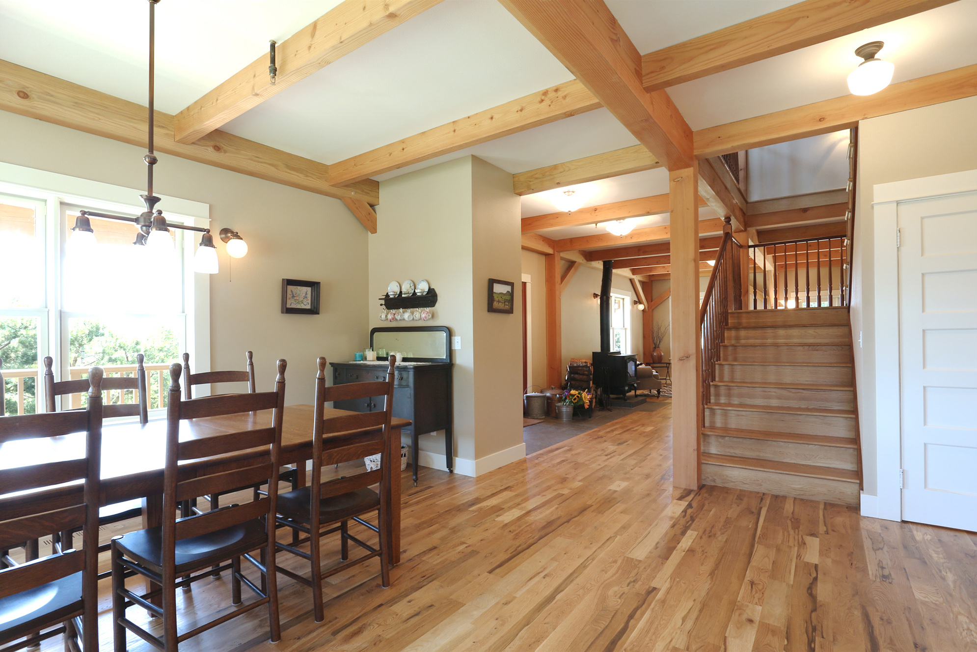 Open plan view from dining table toward central stair and living room with wood stove, exposed timber frame columns connecting all levels, North Plains Timber Frame by Tweto Architecture