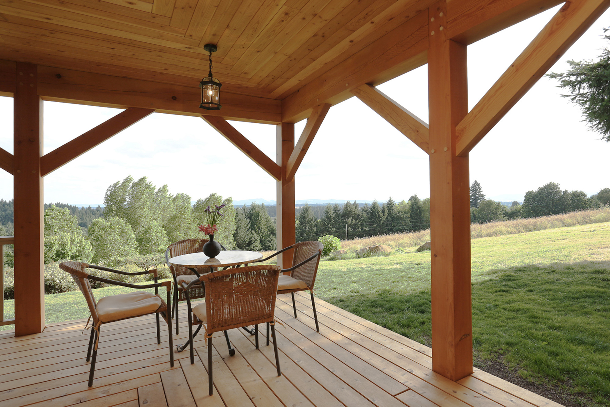 Covered south porch with exposed timber columns and knee braces, outdoor dining table, and sweeping views across rural hillside, North Plains Timber Frame by Tweto Architecture