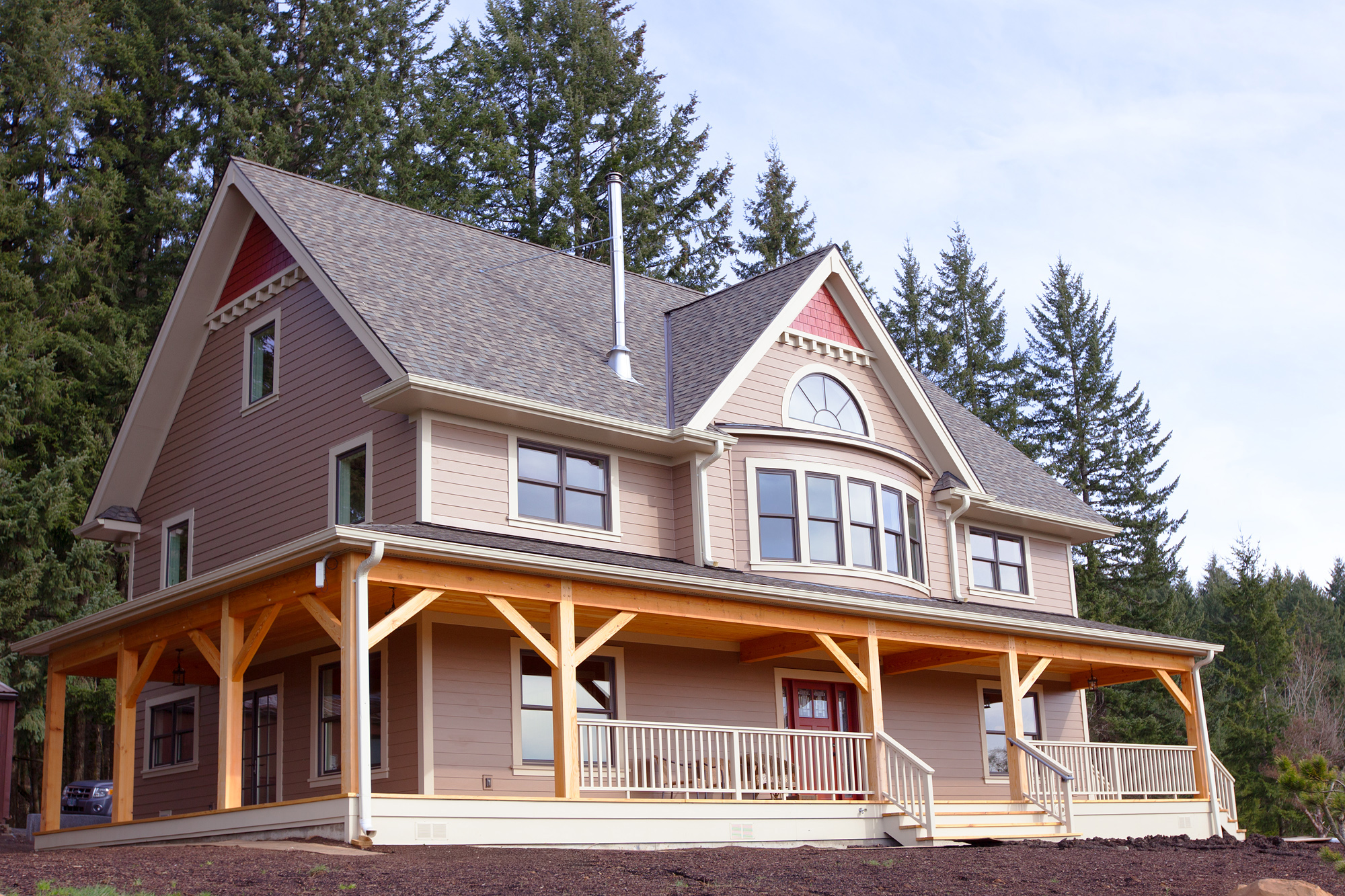 South elevation of completed home with wraparound timber porch, farmhouse gable with half-round window, and surrounding fir trees, North Plains Timber Frame by Tweto Architecture