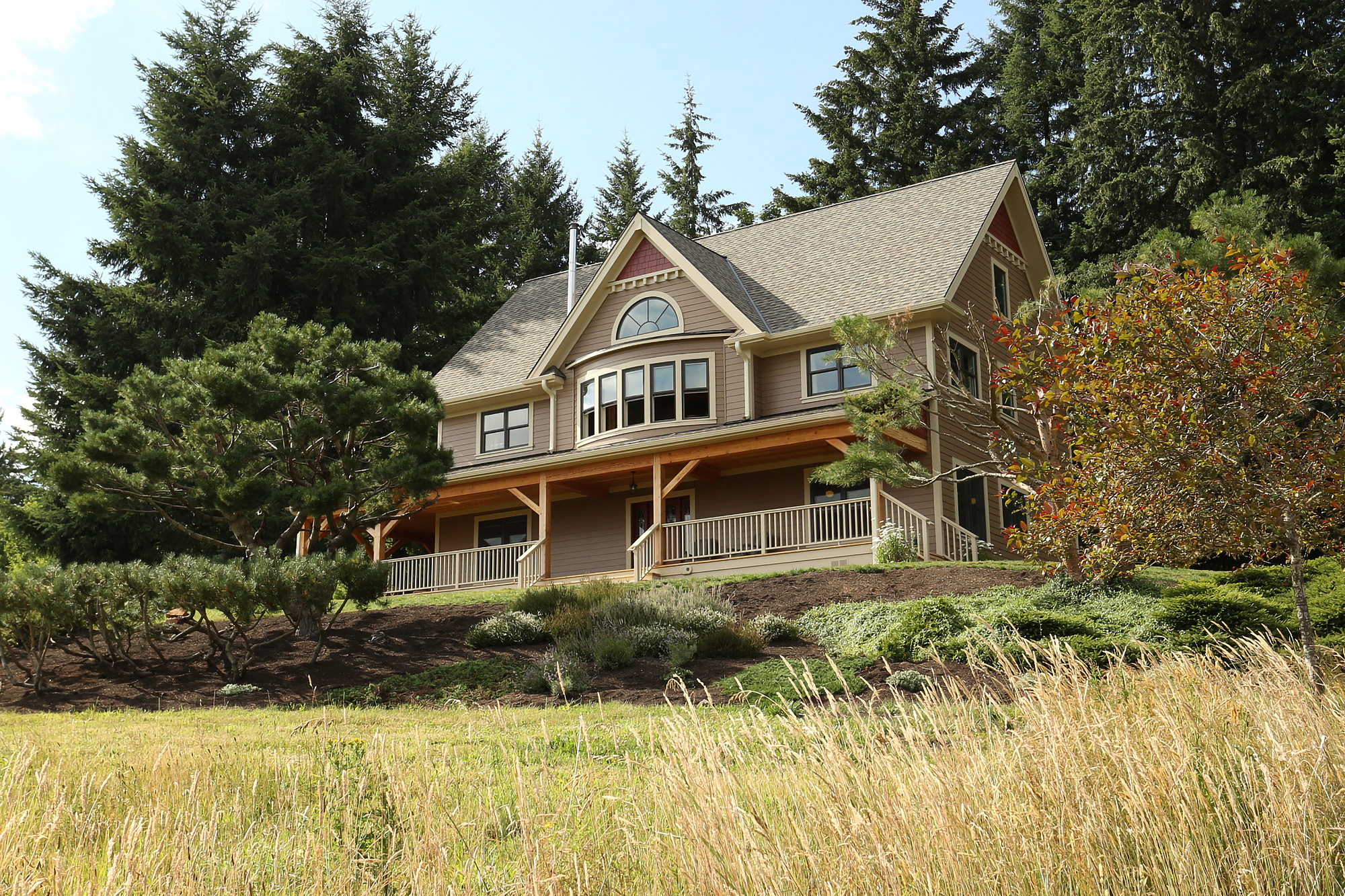Home set into south-facing hillside among mature trees, wraparound porch with exposed timber structure visible from meadow, North Plains Timber Frame by Tweto Architecture