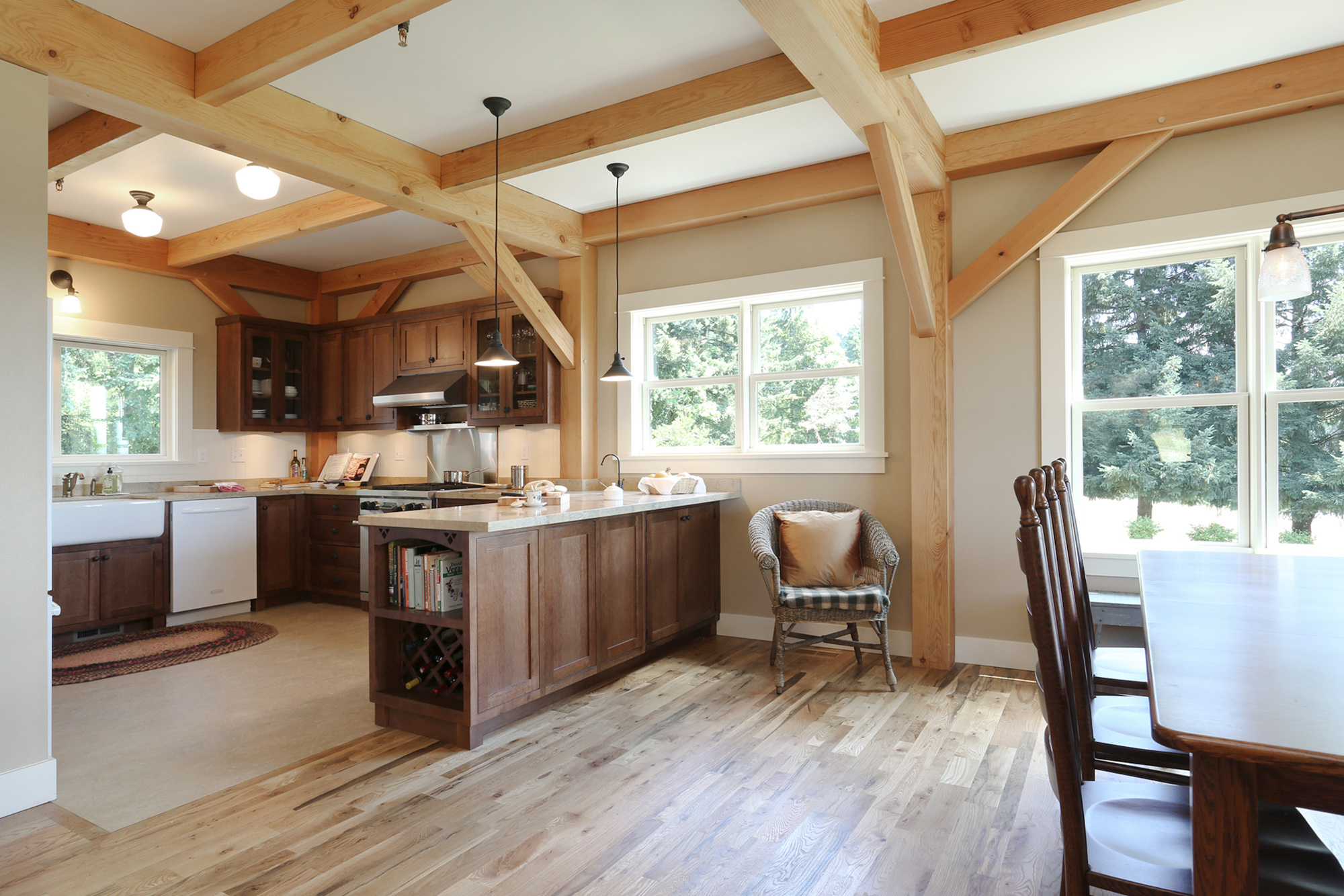 Kitchen from dining side showing walnut island with cookbook shelf and wine rack, farmhouse sink at counter run, exposed timber frame overhead, North Plains Timber Frame by Tweto Architecture
