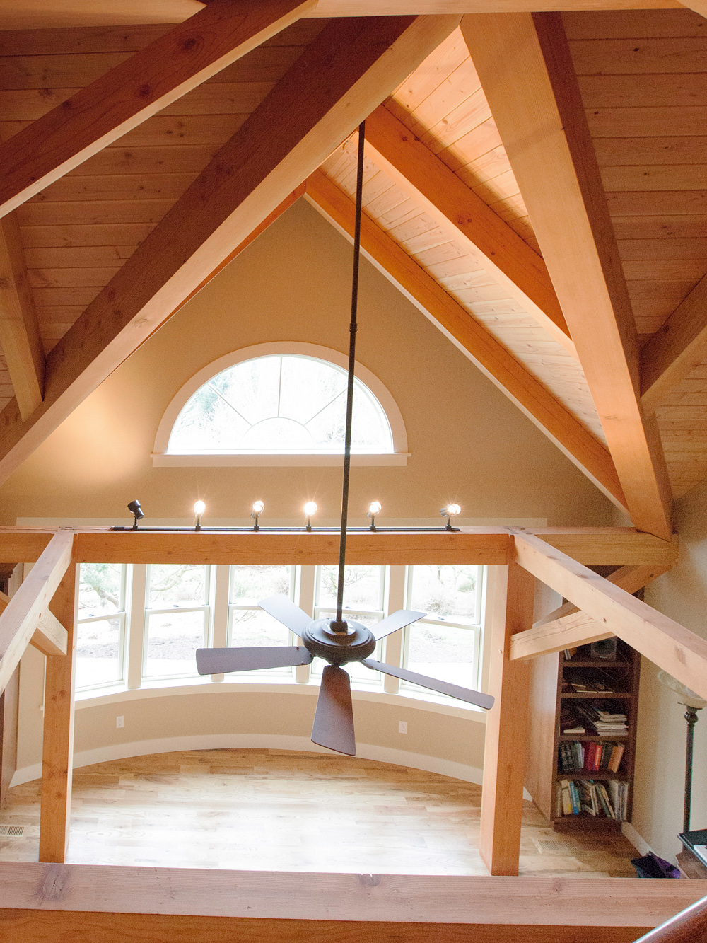 Looking up from loft into vaulted timber frame apex with half-round arched window, ceiling fan pendant, and south-facing windows below, North Plains Timber Frame by Tweto Architecture