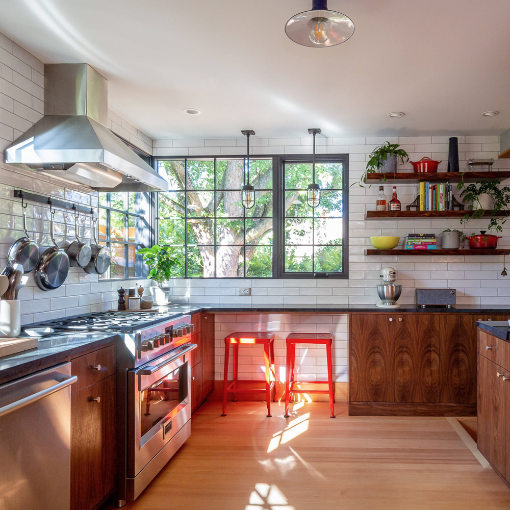 Remodeled kitchen with walnut cabinets, white subway tile, stainless range, large black-framed windows to a mature tree, and open wood shelving, by Tweto Architecture